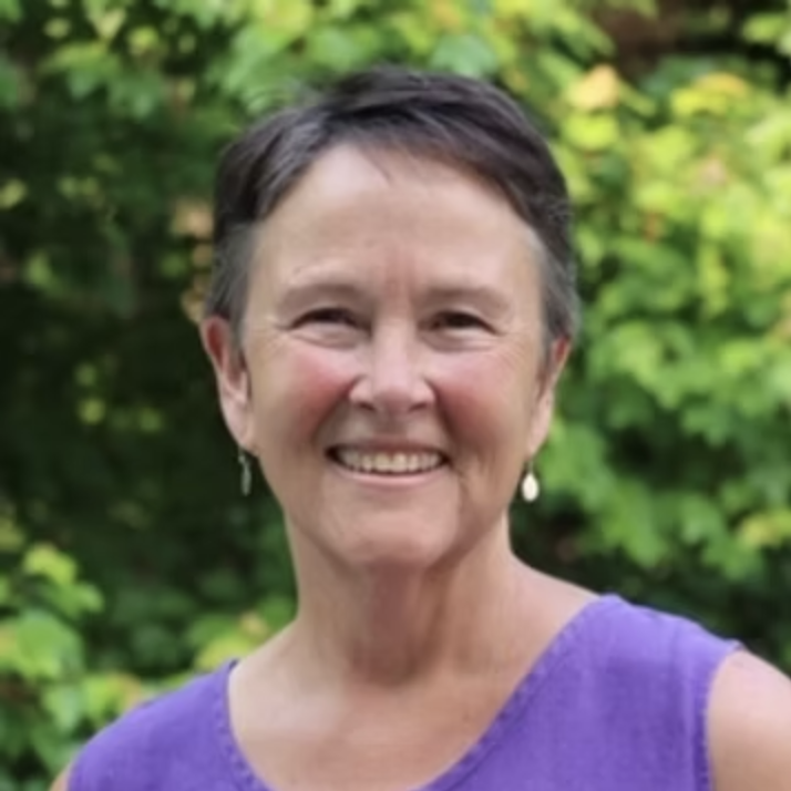 Photo of Sarah, a white woman with short brown hair in her 60s, wearing a purple shirt and earrings.